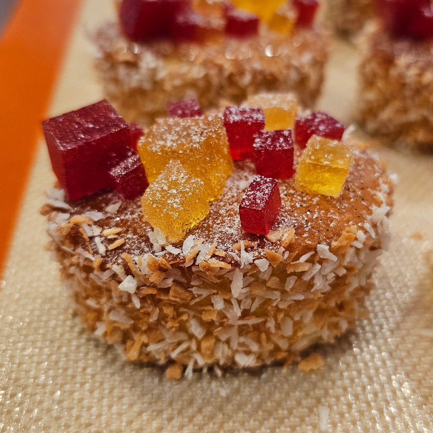 Oval tea cake coated in coconut flakes and topped with cubes of raspberry and lemon pate de fruit.