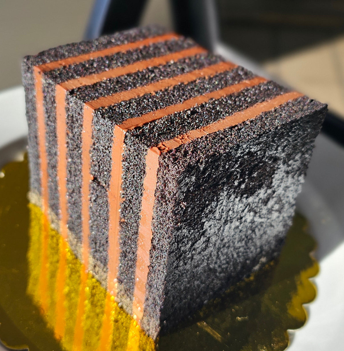 A square layered cake with alternating dark chocolate cake and brown layers of chocolate cremeux, standing on a gold-colored scalloped cake board on a white plate.