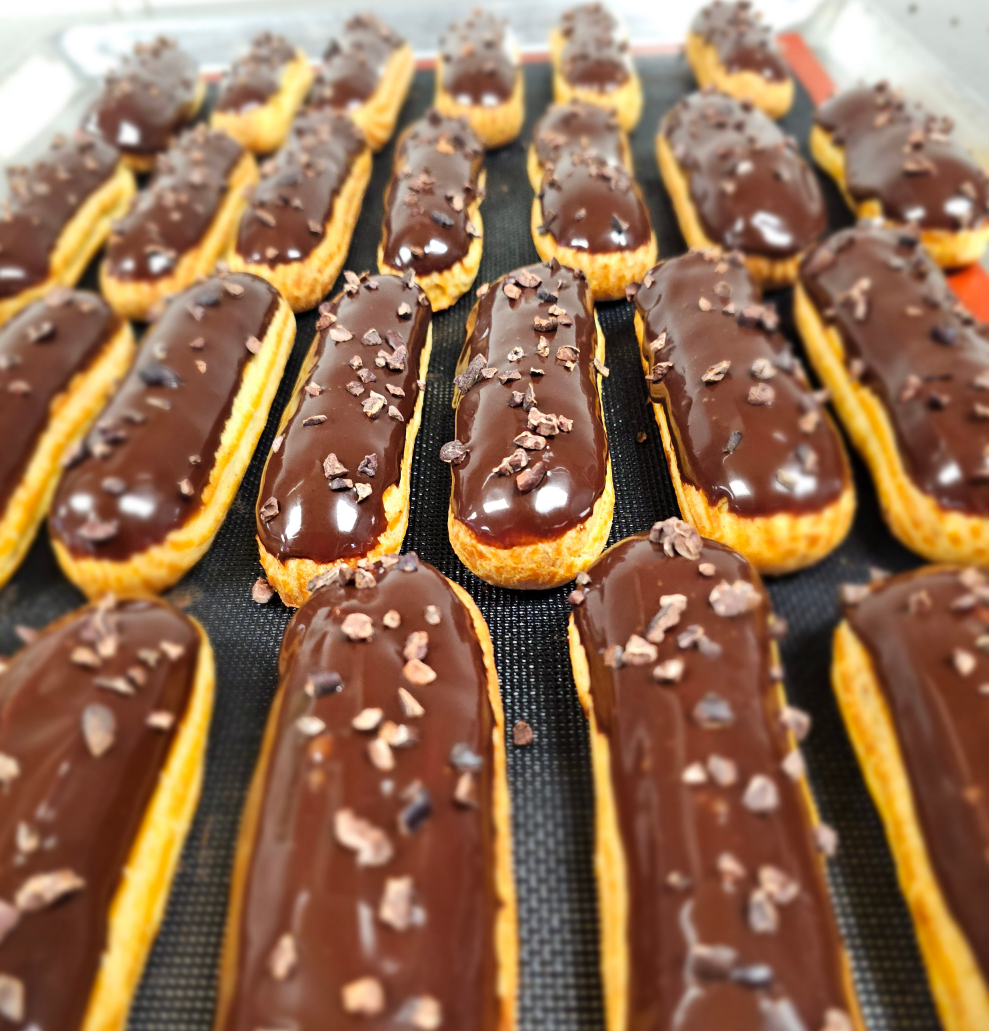 Several chocolate eclairs lined up. Each one coated in chocolate and sprinkled with cacao nibs.