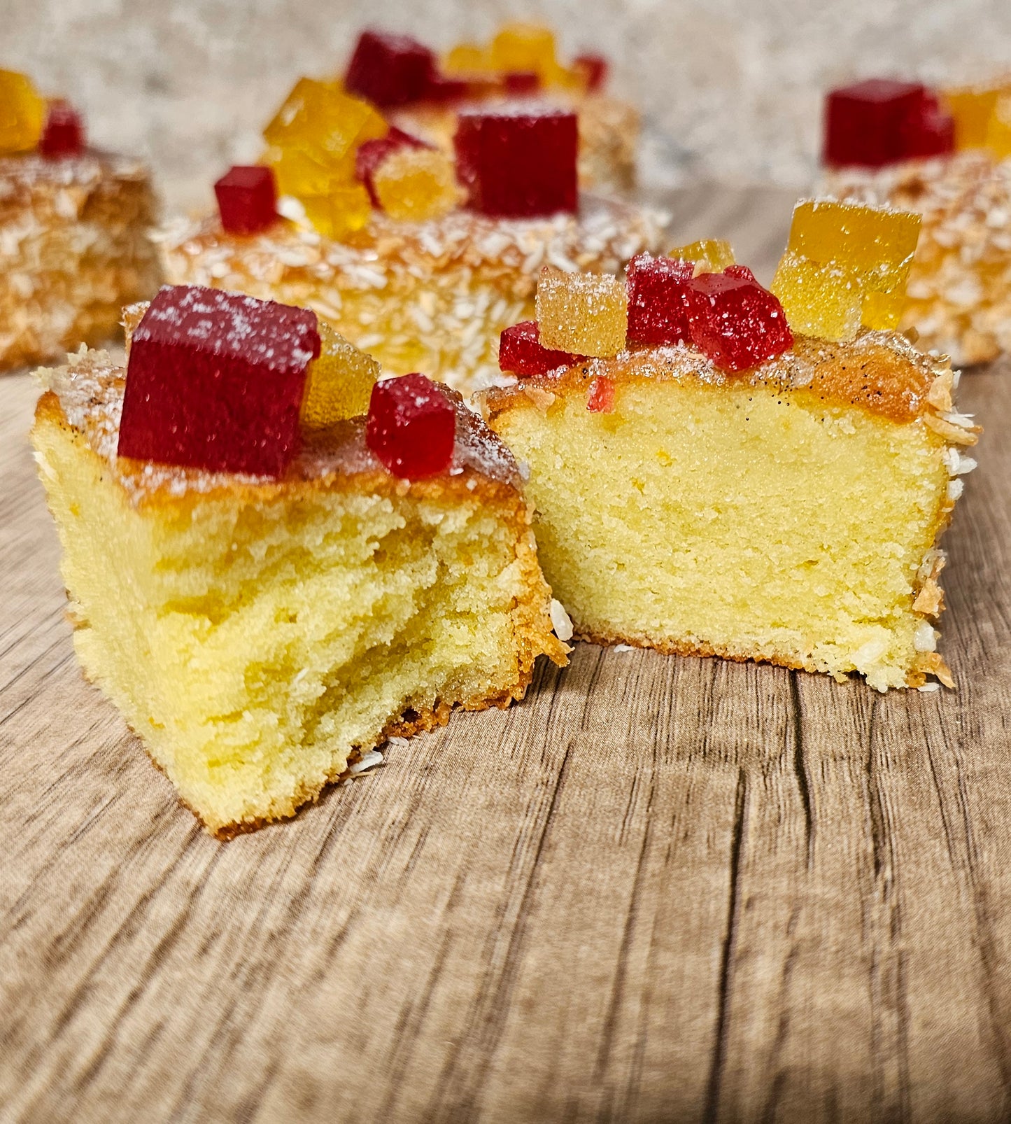 Oval tea cake coated in coconut flakes and topped with cubes of raspberry and lemon pate de fruit. Cake has been sliced in half to show texture.