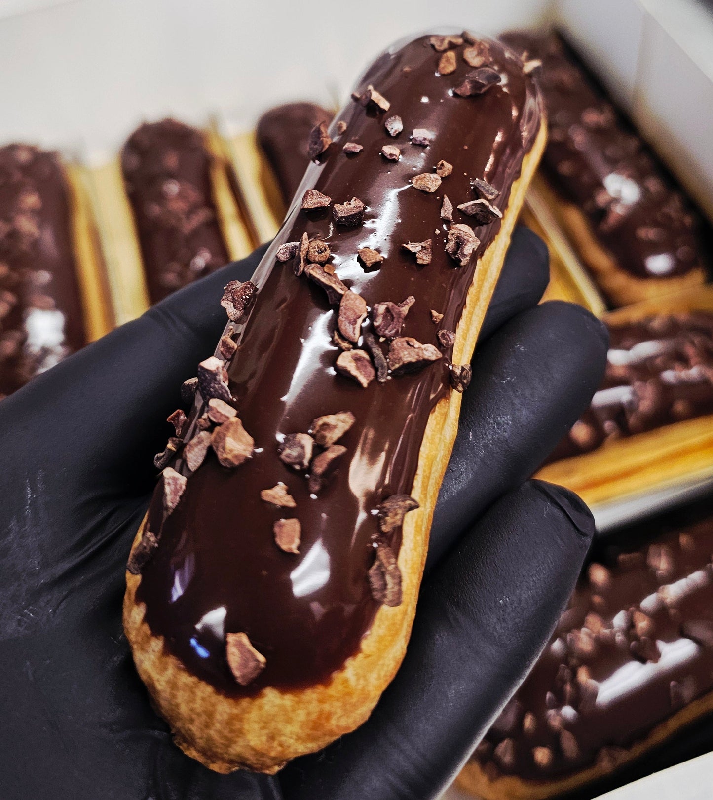 Chocolate-covered eclair with cocoa nibs held by a gloved hand.