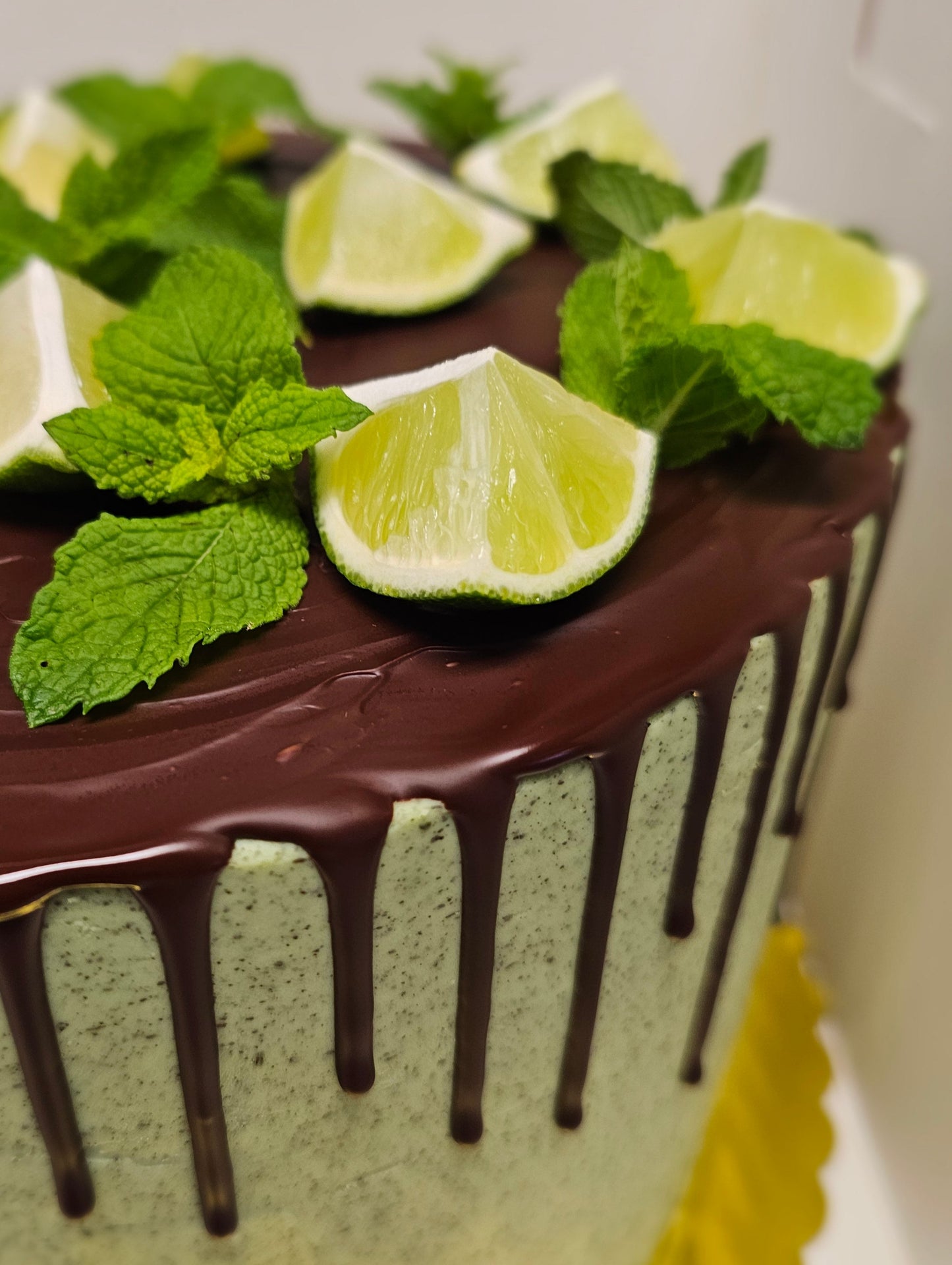 Closeup of a tall mint green frosted cake topped with chocolate drips, lime wedges and fresh mint leaves.