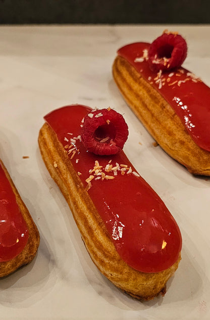A photo showing three coconut raspberry eclairs with a glossy red icing and a raspberry on top, placed on a white surface.