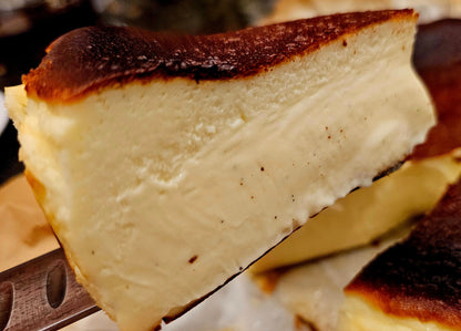A close-up of a slice of basque cheesecake being lifted on a metal spatula, showing a smooth creamy interior and a dark caramelized top crust, with more cake visible in the background on parchment paper.