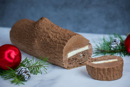 Sliced Buche de Noel mousse cake revealing layer inside. Pine branches and red balls used for decoration against a blue background