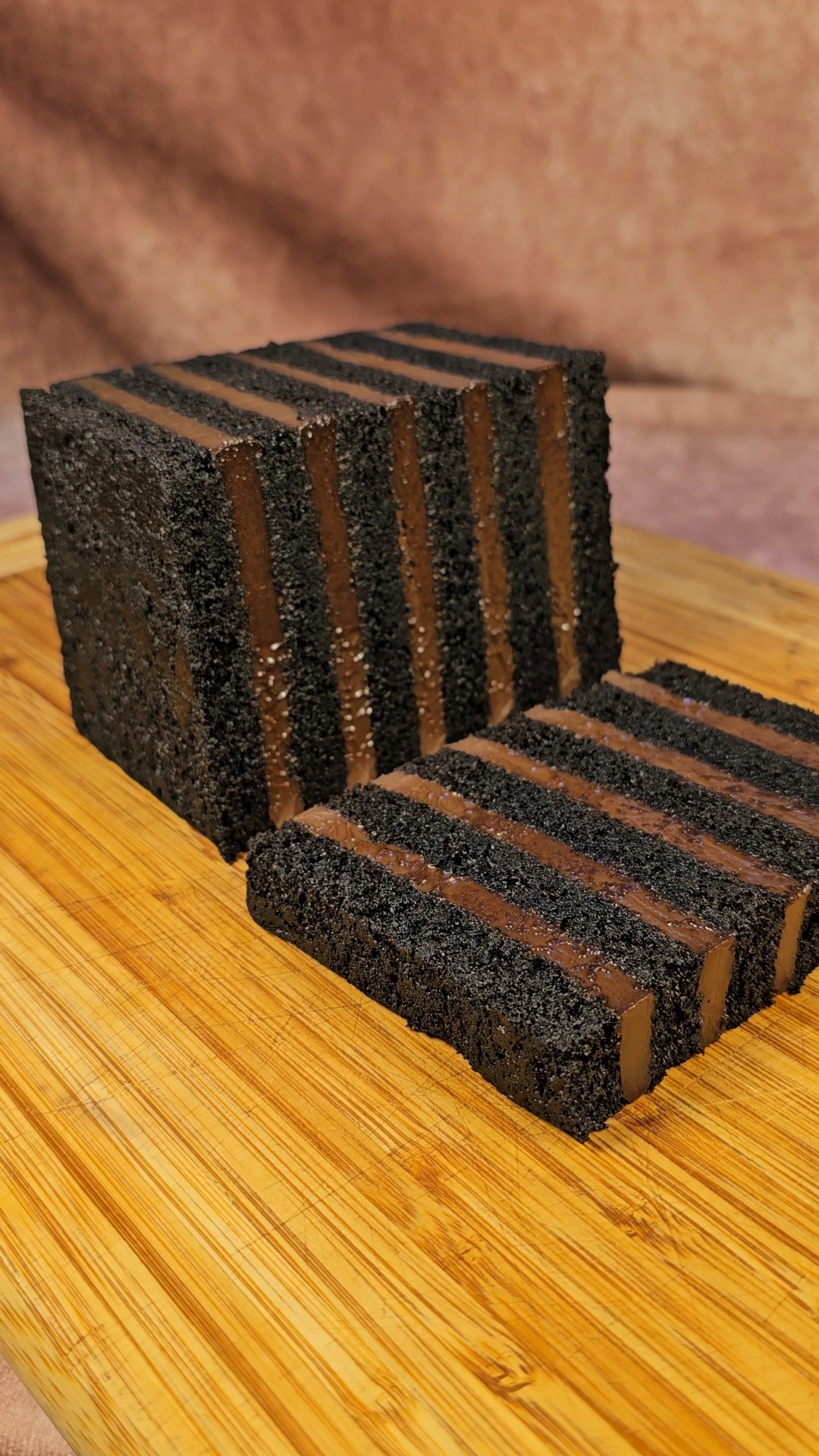 A rectangular dark chocolate layer cake with alternating dark chocolate cremeux layers, shown sliced and standing on a wooden board against a warm brown background.