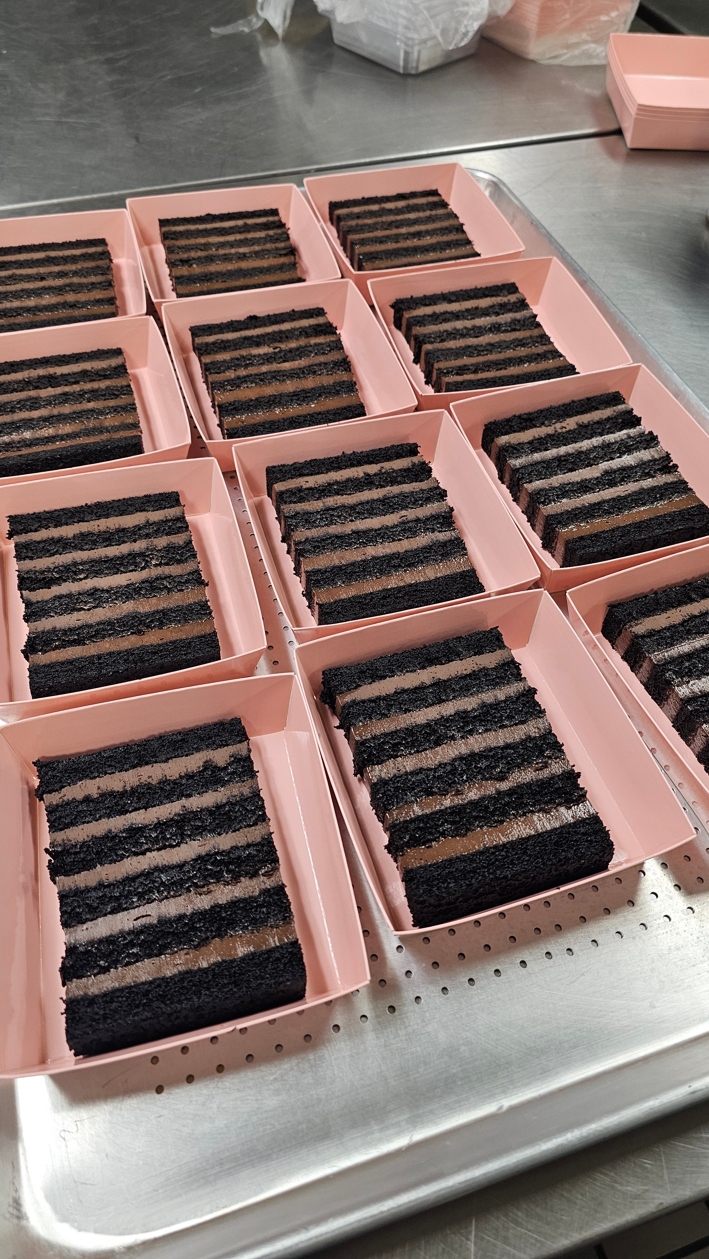 Multiple rectangular slices of dark chocolate layer cakes in pink boxes  arranged on a metal tray.