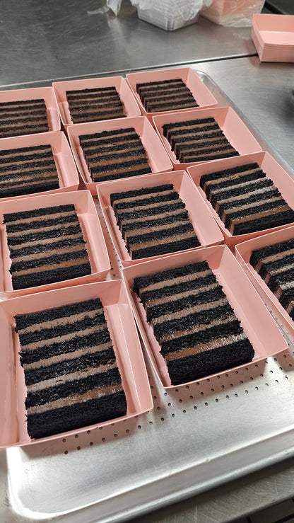 Multiple rectangular slices of dark chocolate layer cakes in pink boxes  arranged on a metal tray.