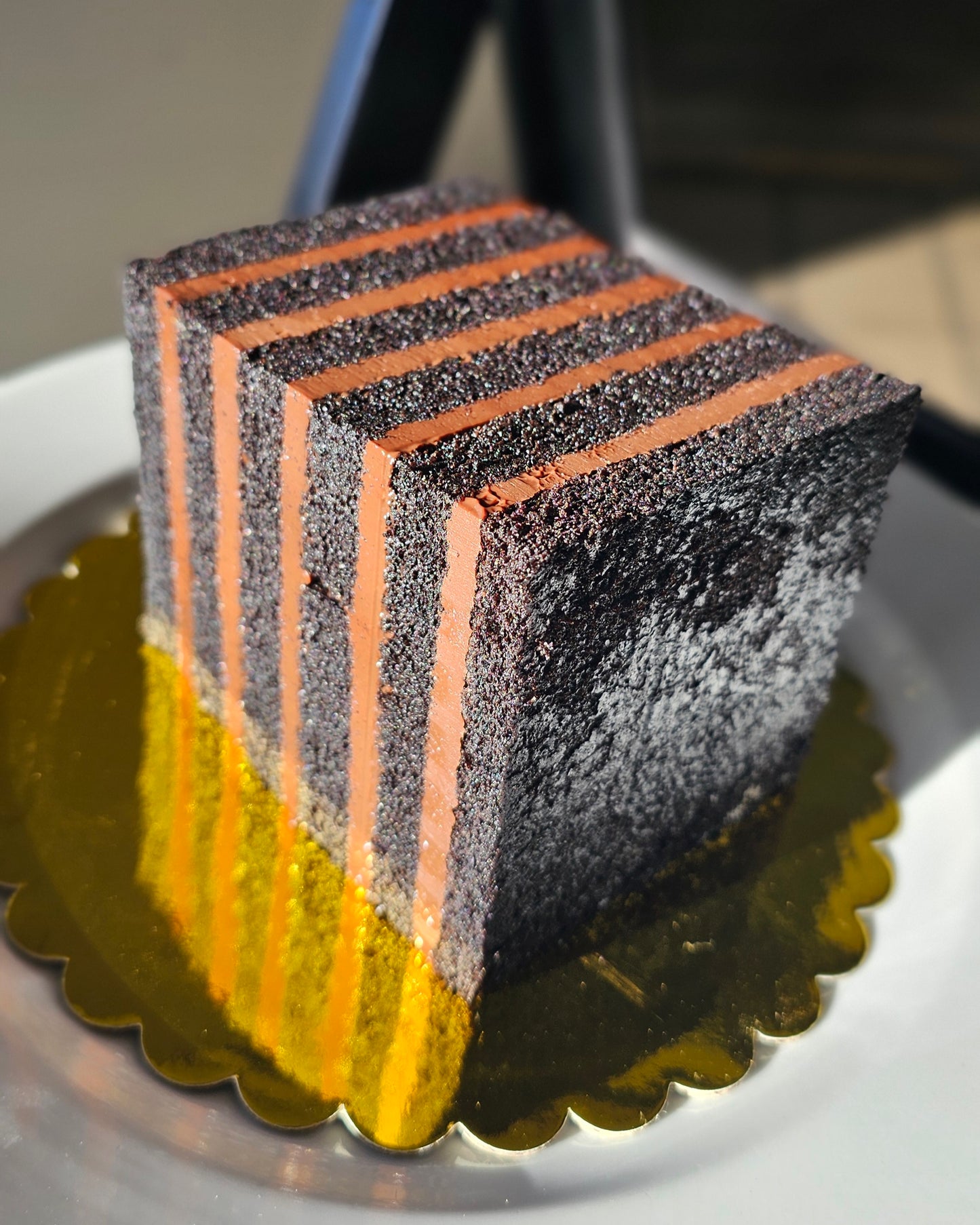 A square layered cake with alternating dark chocolate cake and brown layers of chocolate cremeux, standing on a gold-colored scalloped cake board on a white plate.