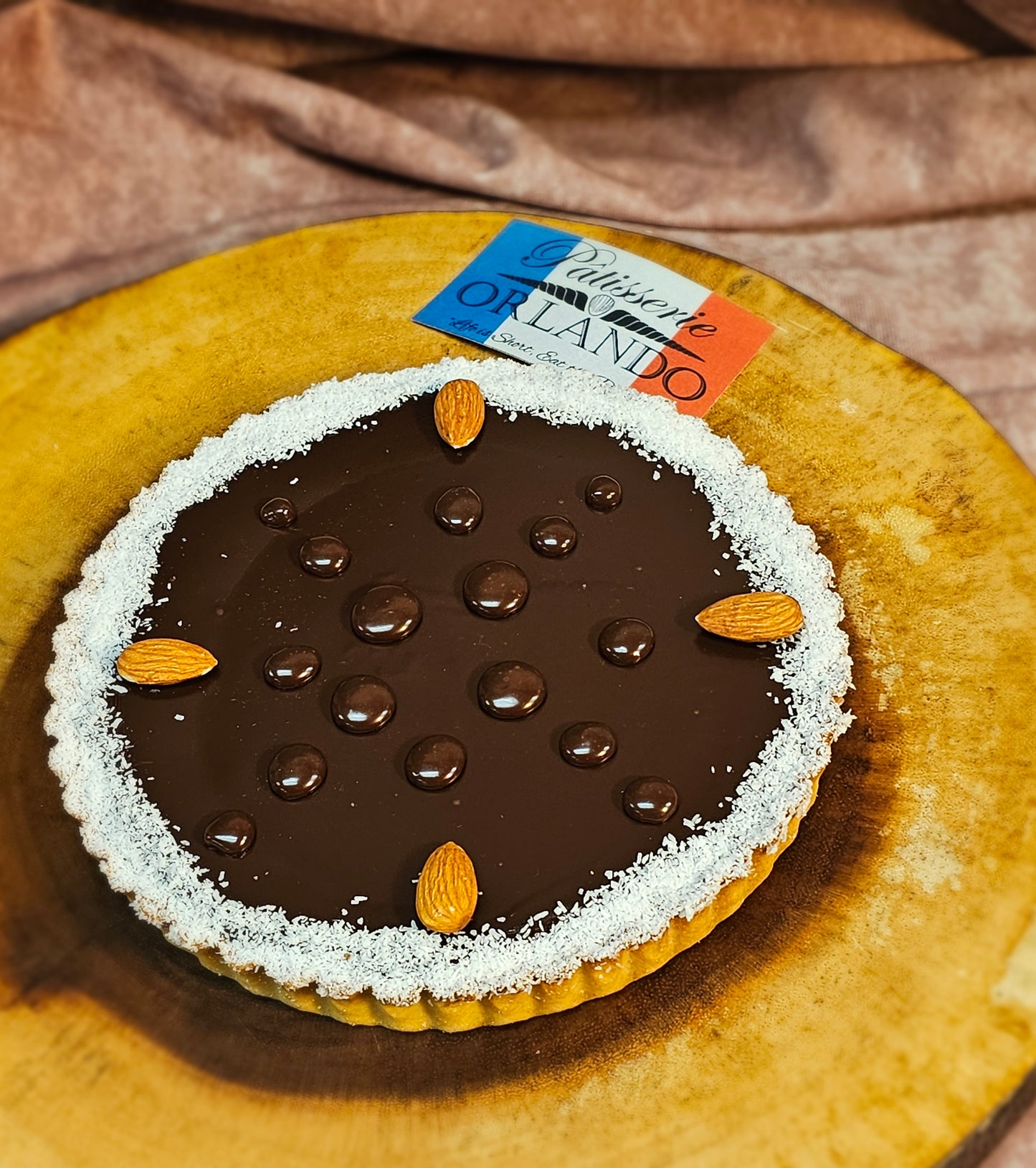 Chocolate tart with almonds on a wooden board, featuring a 'Patisserie Orlando' label.