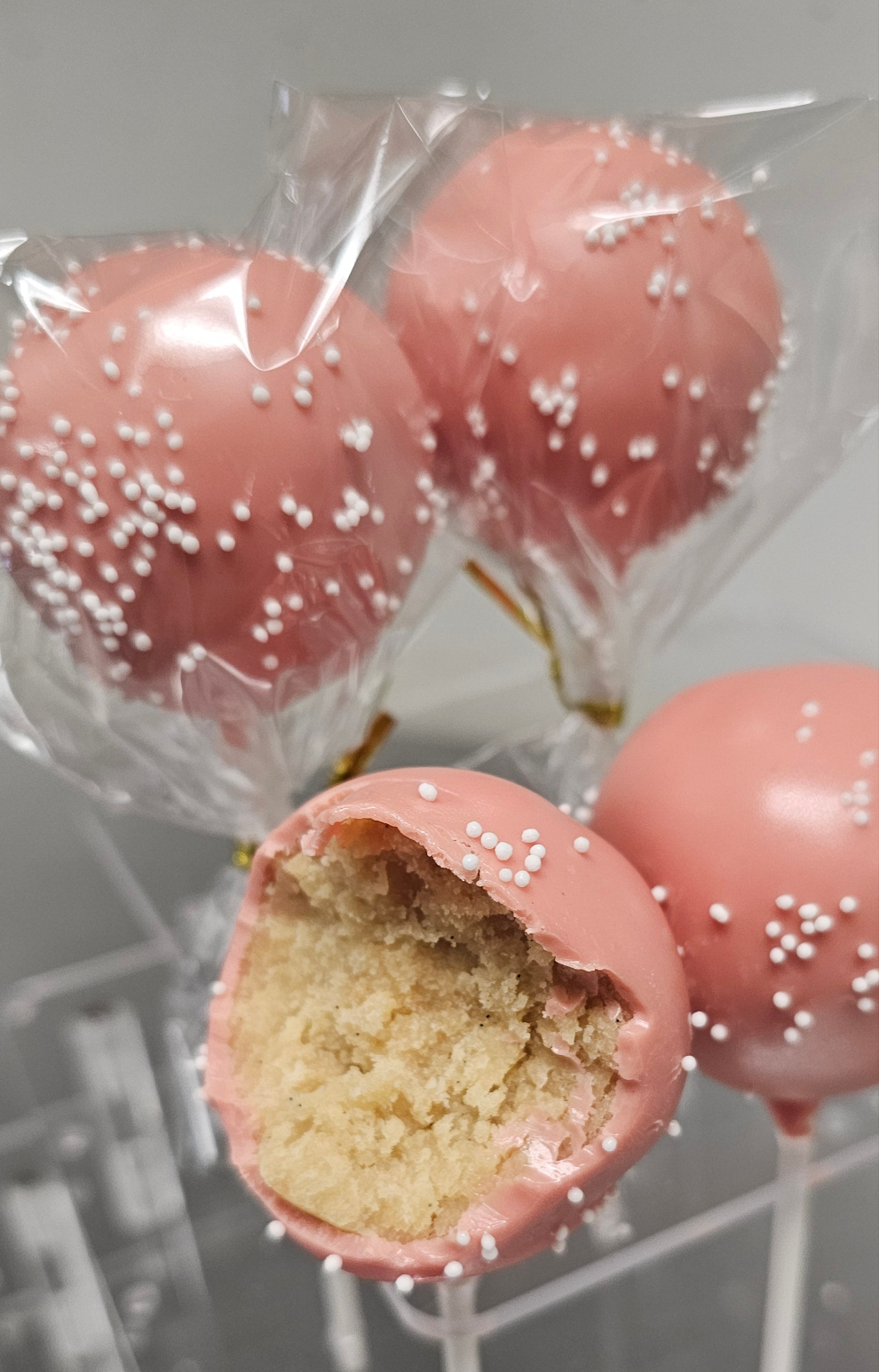 Four pink, round cake pop with white sprinkles. One has a bite taken from it.