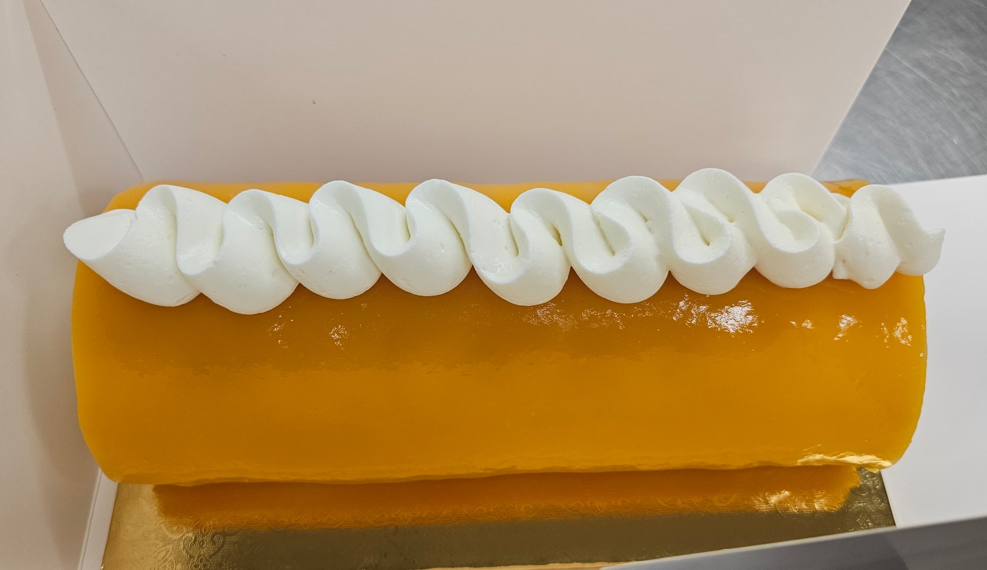 Roll cake coated with orange mango glaze and white S shaped cream across the top.