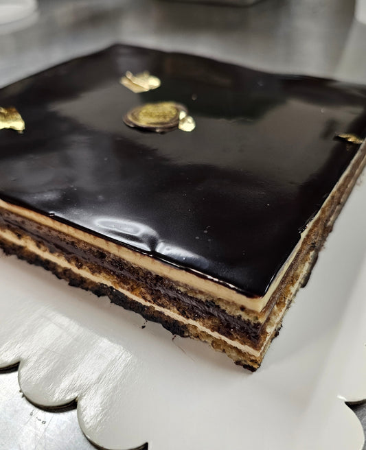 Opera cake with a shiny chocolate glaze on top, showing multi layered sides, sitting on a white board. 