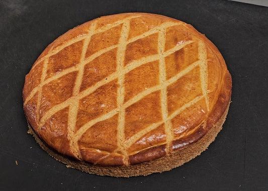A round golden-brown baked bread or pastry with a diamond lattice pattern cut on top, resting on a dark gray surface.