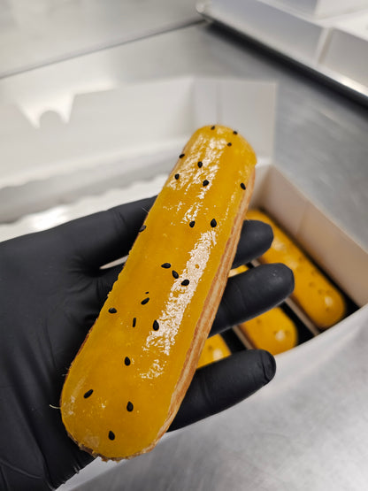 Person wearing black gloves holding an eclair with yellow glaze and black seeds.