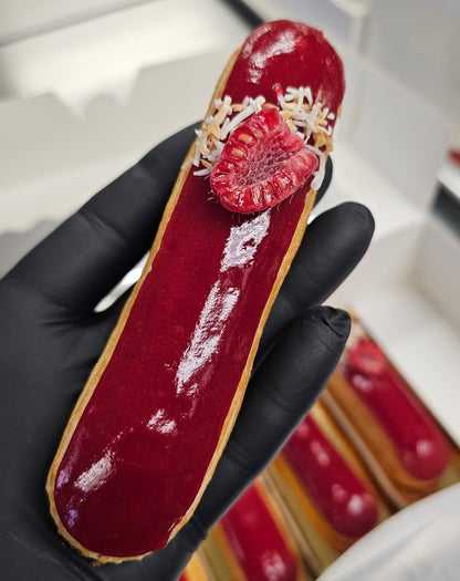 A black gloved hand holding an eclair with a deep red glaze and decorated with a cut raspberry and shredded coconut.