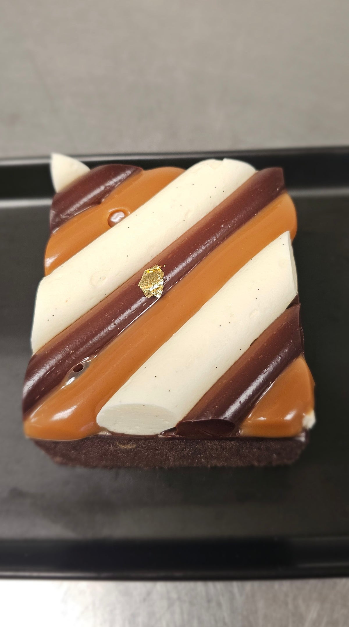 A square chocolate cake slice topped with diagonal stripes of white cream, caramel sauce, and dark chocolate, with a small gold leaf decoration, resting on a black plate.