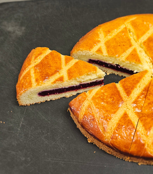 A golden-brown baked cake with a diamond crisscross pattern on top and a dark berry jam filling, shown with one slice cut and set beside the main round cake on a dark slate surface.