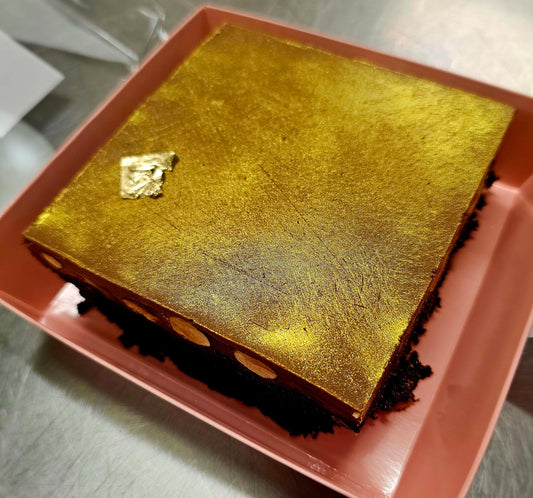 A square brownie dusted with gold powder and a small gold leaf accent, presented in a pink square container on a stainless steel surface.