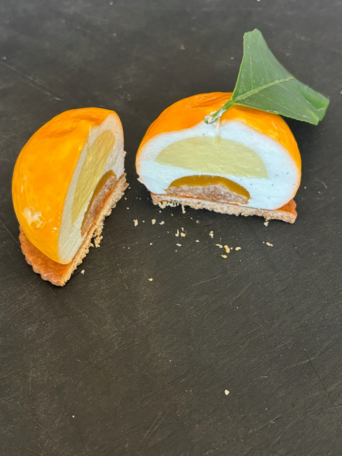 Individual mousse cake shaped like a mandarin, sliced to reveal three layers inside. A cake layer, a couli layer and a custard layer.